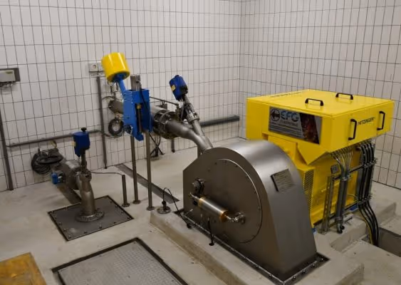 Industrial room with a yellow generator and various metal pipes and valves mounted on a concrete floor against tiled walls.