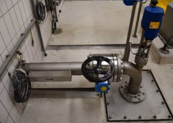 Industrial metal pipe with a black circular valve wheel and attached blue control device in a tiled utility room.