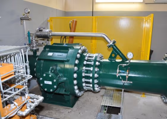 Industrial green mechanical pump with pressure gauges and metal pipes inside a facility with yellow safety screens in the background.