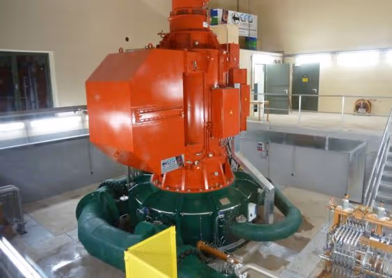 Large industrial turbine with orange and green components inside a mechanical room with concrete walls and metal railings.