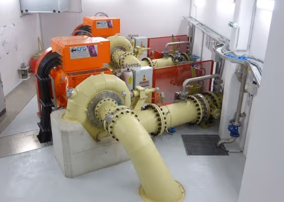 Indoor mechanical room featuring large industrial pipes and orange generator units connected by metal flanges.