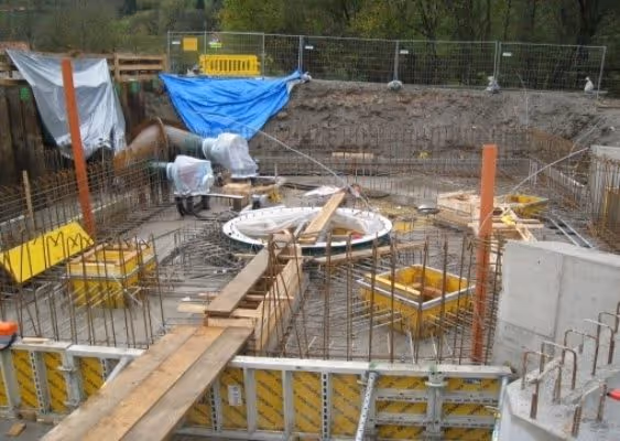 Construction site with concrete foundation work, wooden planks, metal rebar, and yellow formwork panels under a blue tarp.