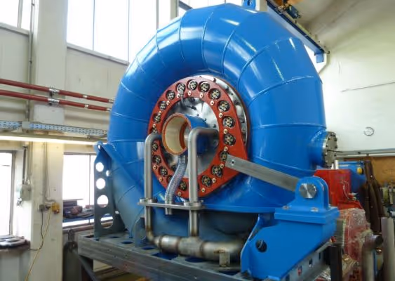 Large blue industrial turbine or generator component inside a workshop with pipes and mechanical parts.