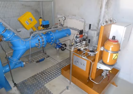 Industrial setup featuring blue pipeline components connected to an orange hydraulic unit with gauges and a yellow control box mounted on a wall.