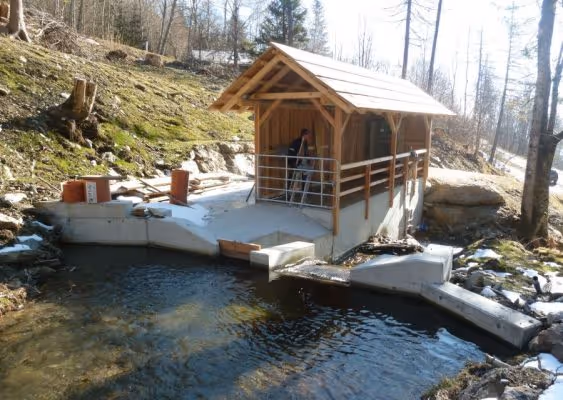 Small wooden structure with a slanted roof on a concrete platform over a clear water stream in a wooded area.