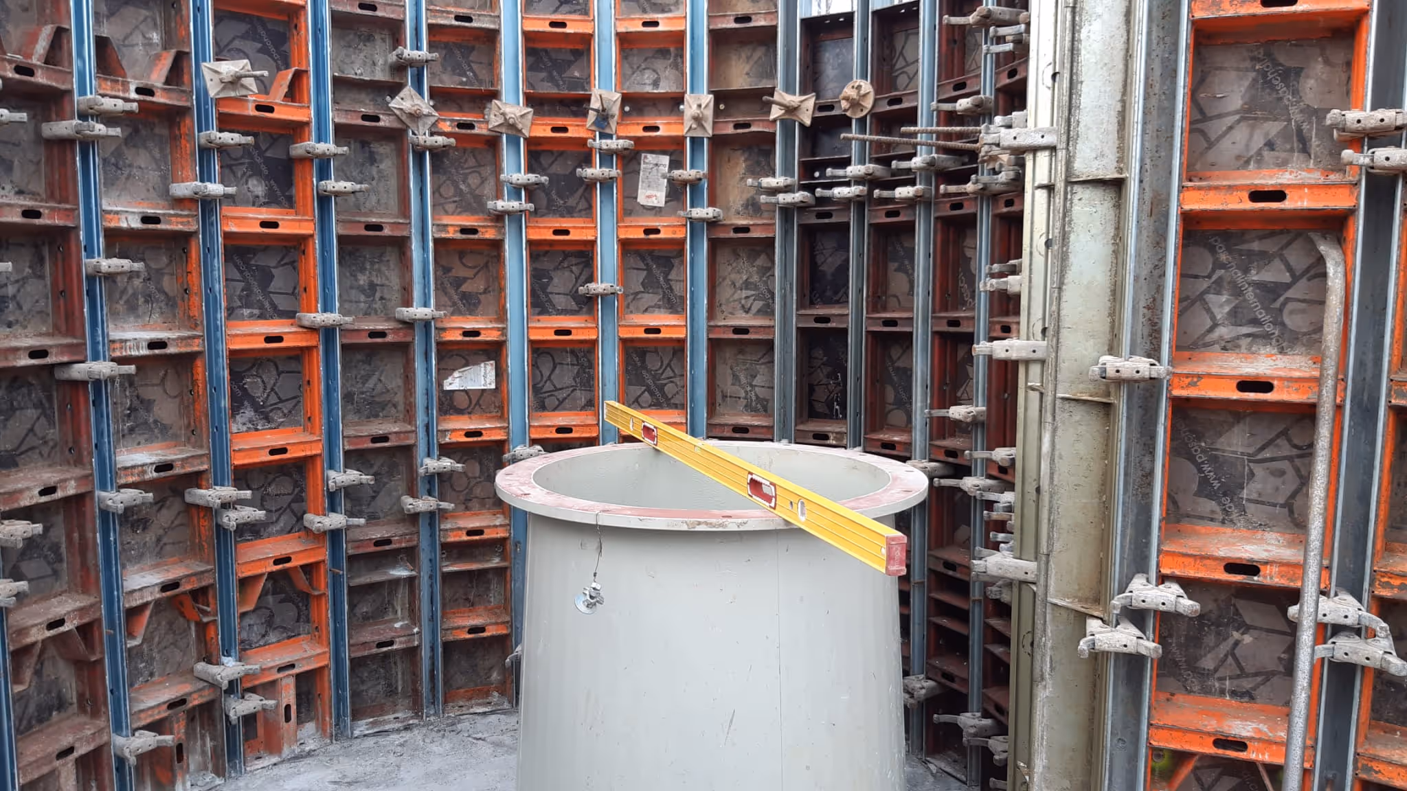 Construction site showing curved concrete formwork with a round concrete pipe in the center and a yellow level tool on top.