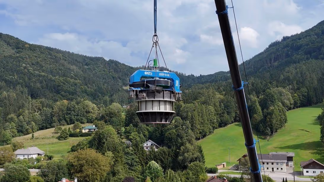 Large mechanical device being lifted by a crane over a green valley with scattered houses and forested hills under a partly cloudy sky.