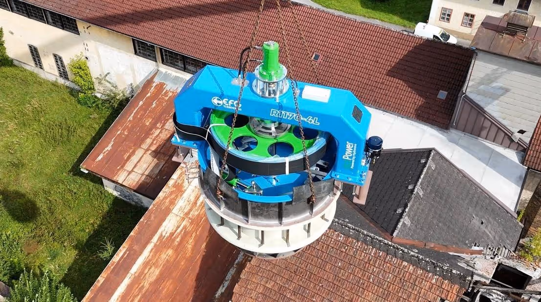 Large blue and green industrial turbine or generator being lifted by chains above rooftops.