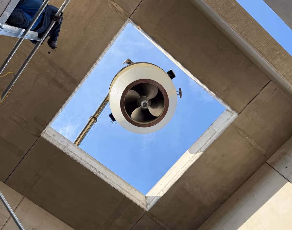 View looking up through a square opening in a concrete structure showing a large industrial fan attached to a pole against a blue sky.