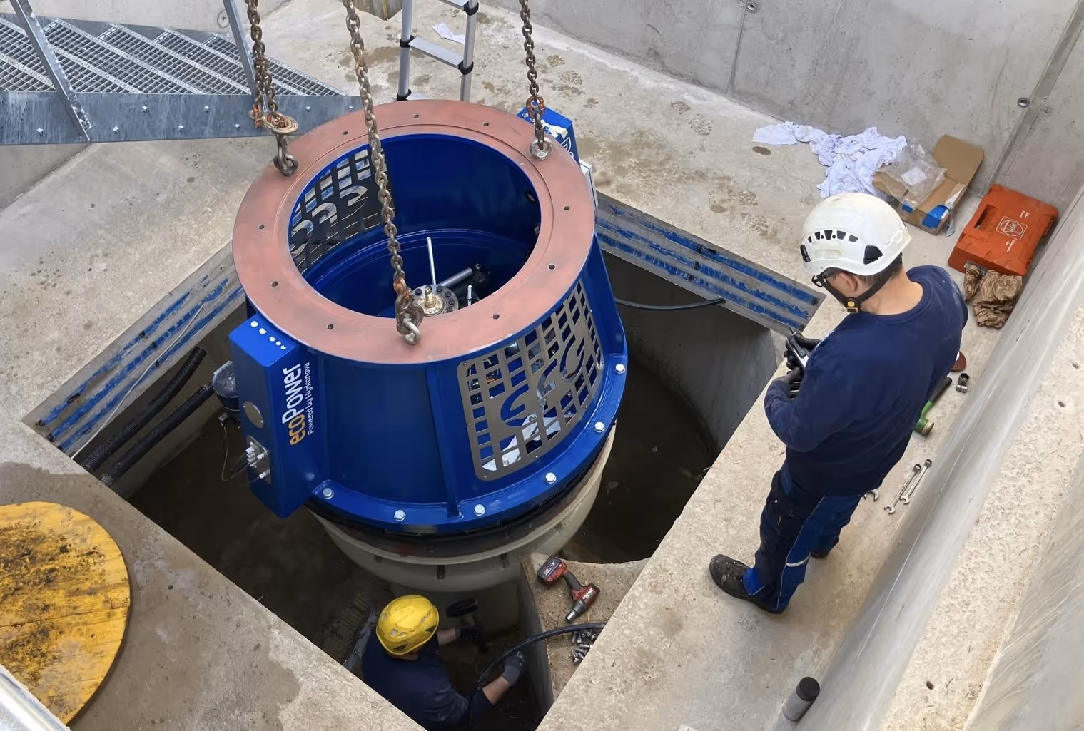 Two workers wearing helmets installing a large blue ecoPower turbine device inside a concrete structure.