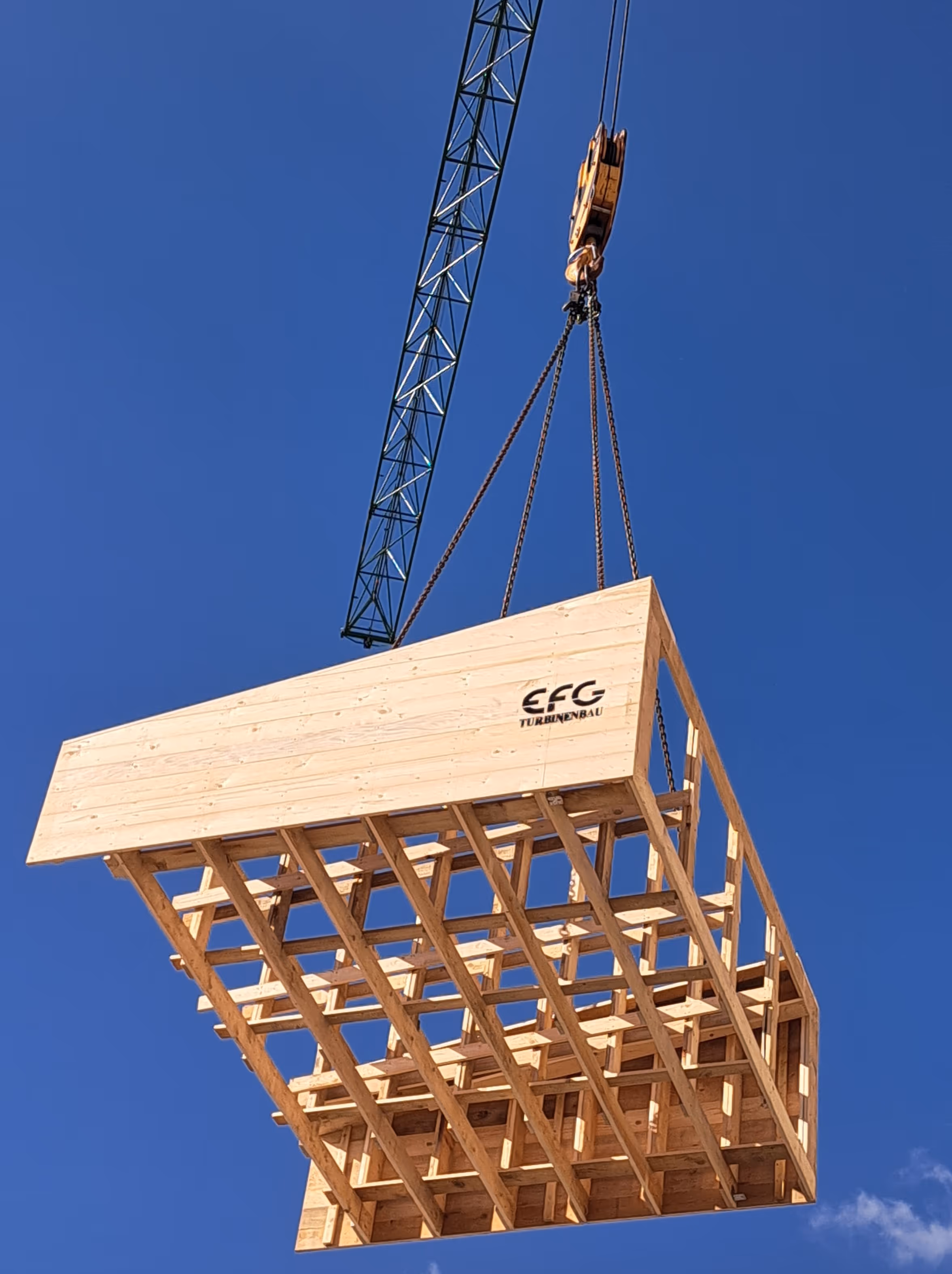 Large wooden structure being lifted by a crane against a clear blue sky.