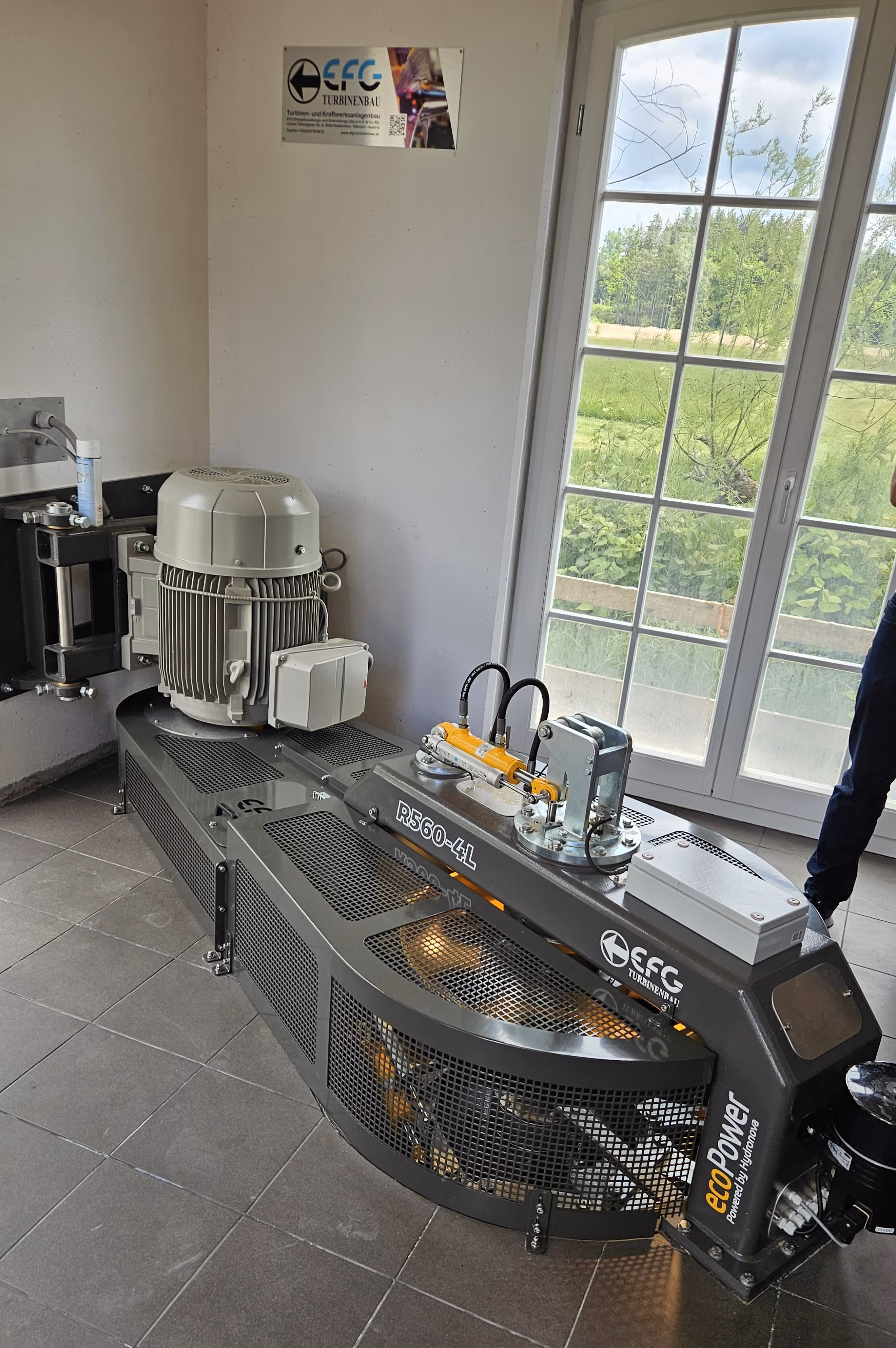 Industrial hydroelectric turbine generator labeled R560-4L installed indoors near a large window with outside greenery visible.