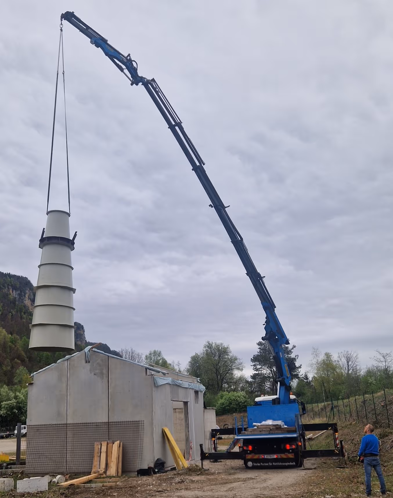 Blue crane truck lifting a large white industrial funnel next to a small concrete building on a cloudy day.