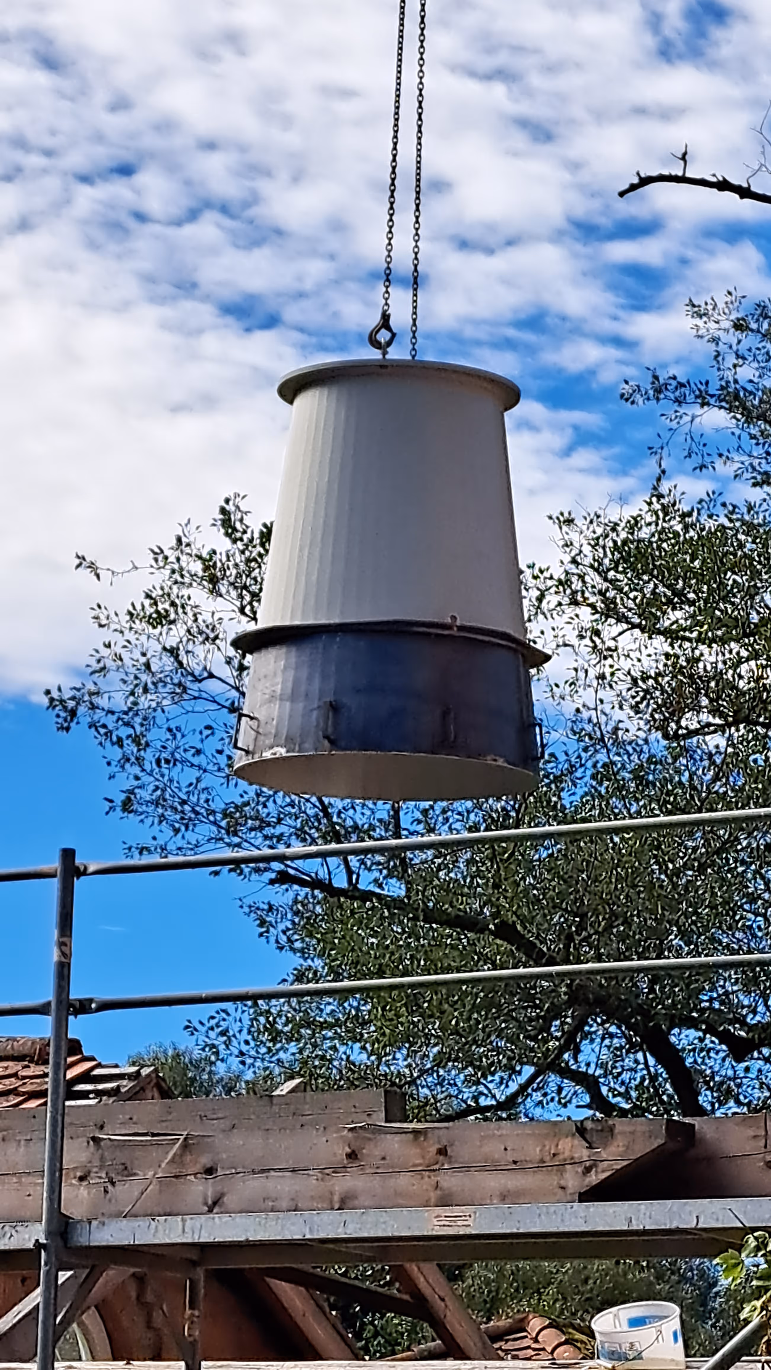 White and gray construction bucket suspended by chains above scaffolding with trees and blue sky background.