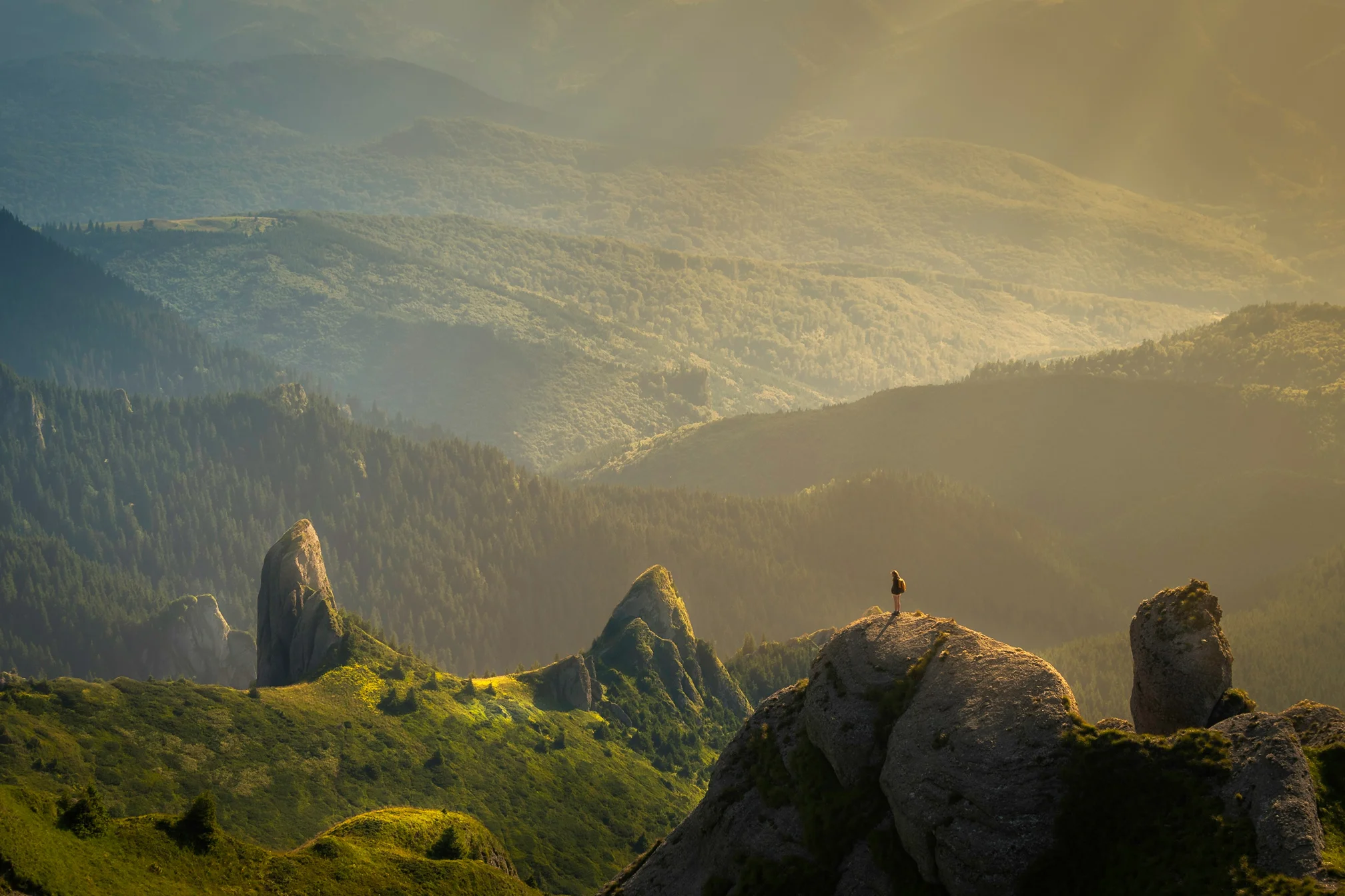 Pessoa em pé no topo de uma rocha olhando para uma paisagem de montanhas verdes iluminadas pelo sol.