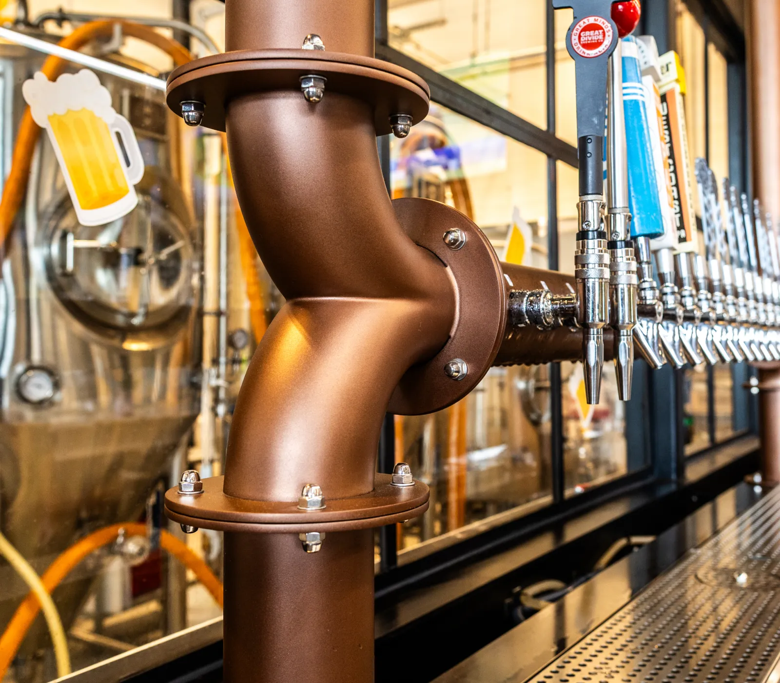 Close-up of beer taps and a large copper pipe in a brewery with brewing equipment in the background.