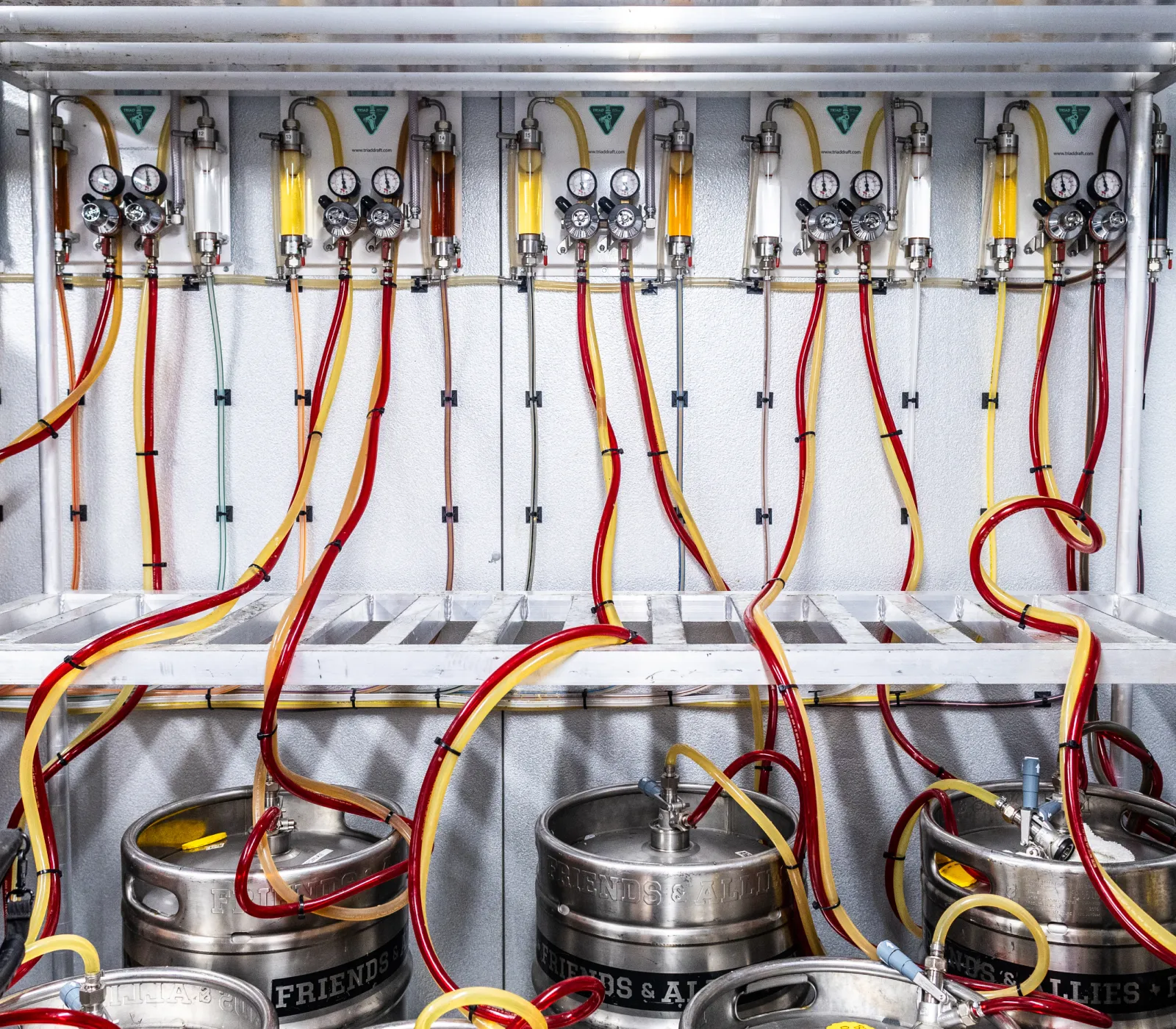 Rows of metal kegs connected to a complex system of gauges and red and yellow tubes in a beverage dispensing setup.