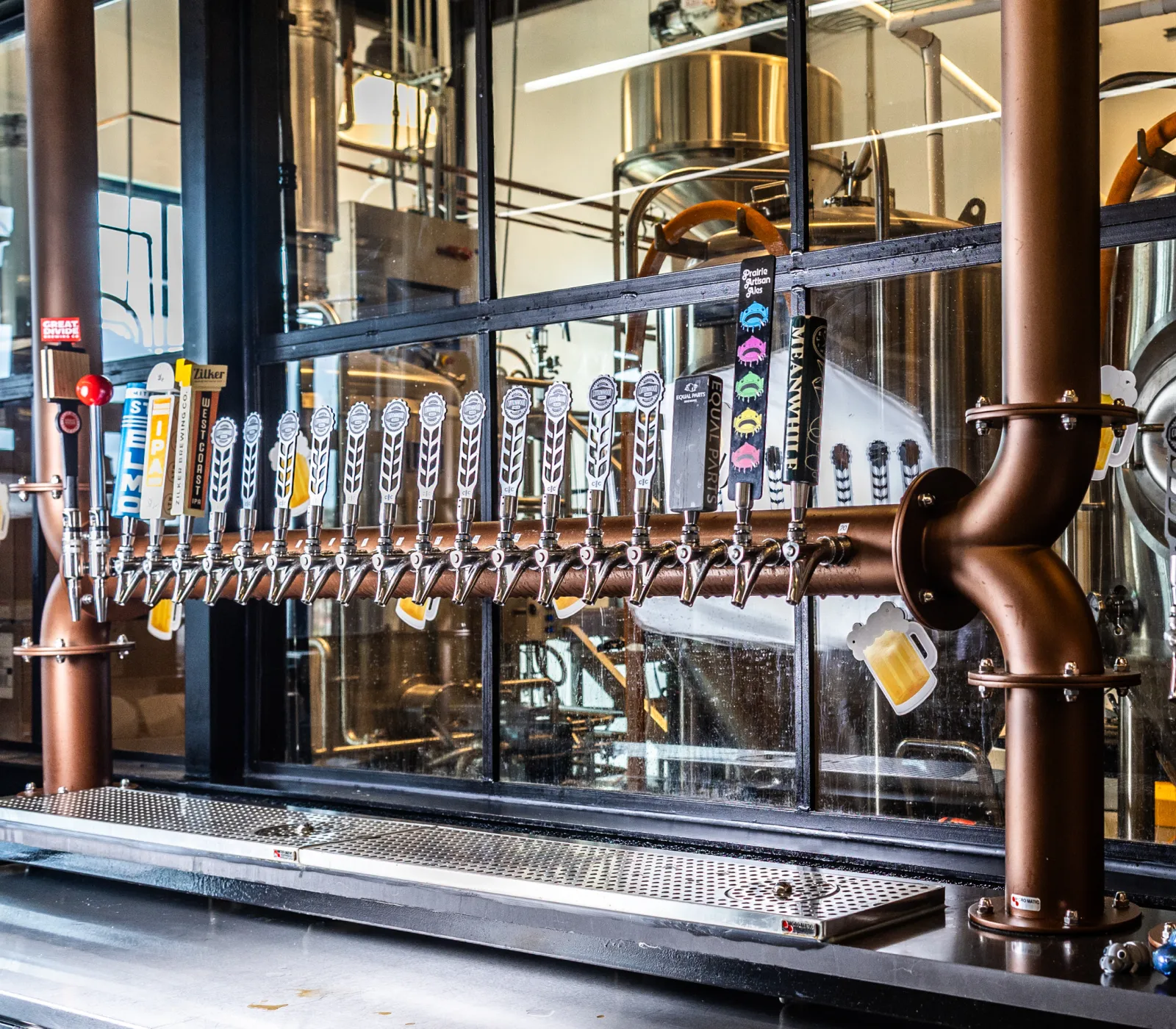 Row of beer taps mounted on a copper pipe in a brewery with brewing equipment visible behind glass.