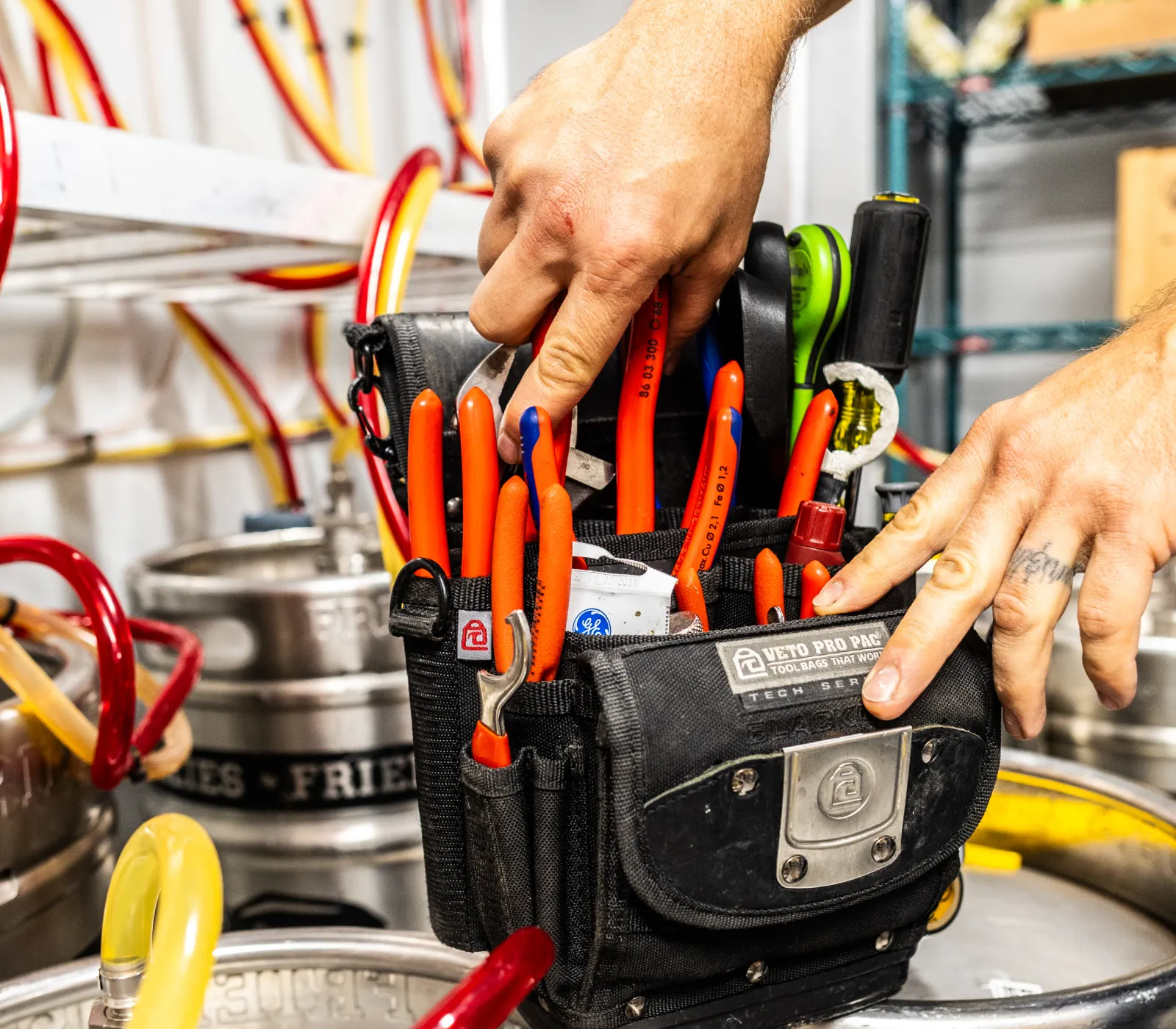 Person's hands reaching into a black Veto Pro Pac tool bag filled with red-handled tools in a workspace with metal kegs and colorful tubing.