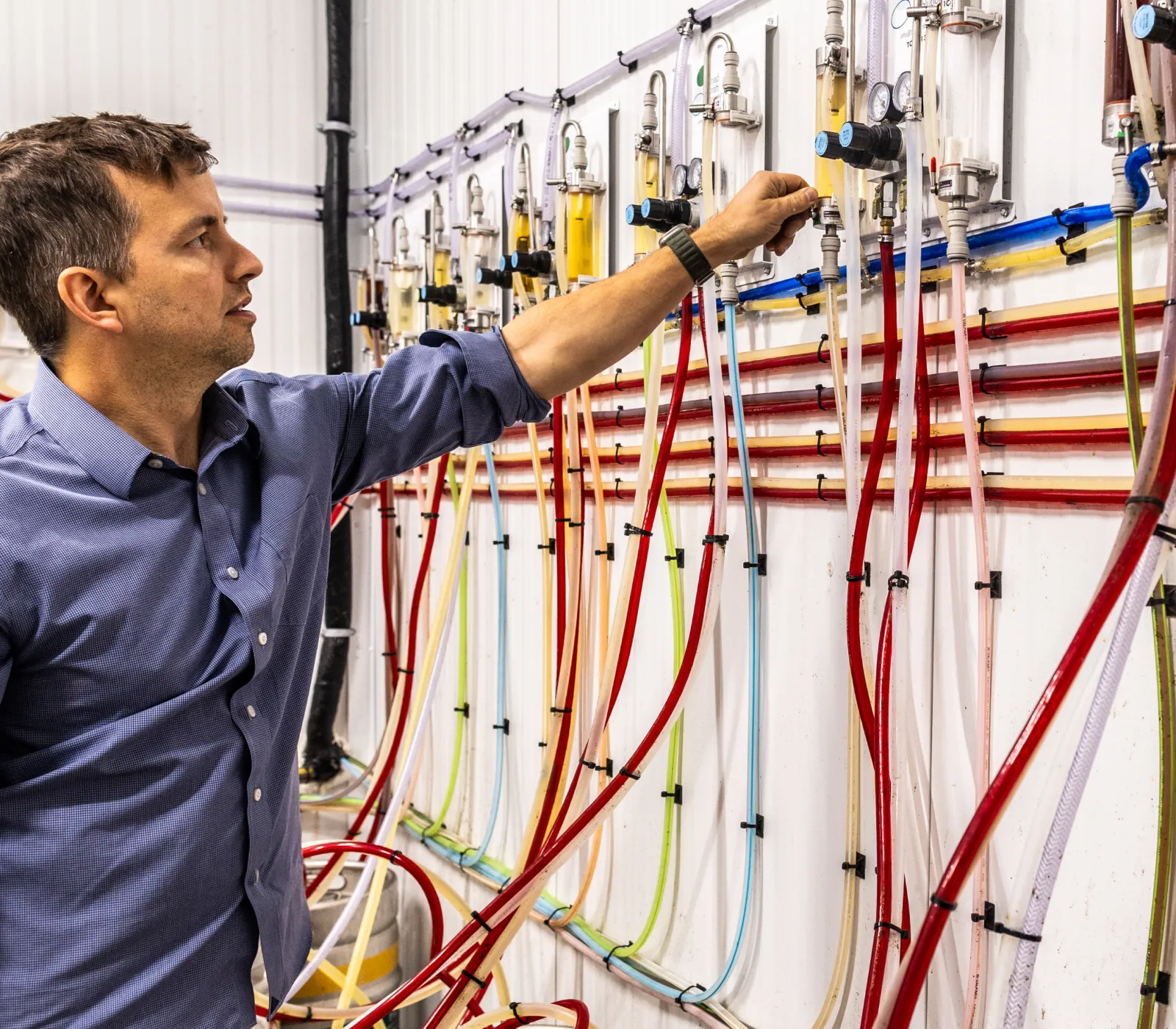 Man in blue shirt adjusting valves on a wall with multiple colorful tubes and flow meters in an industrial or laboratory setting.