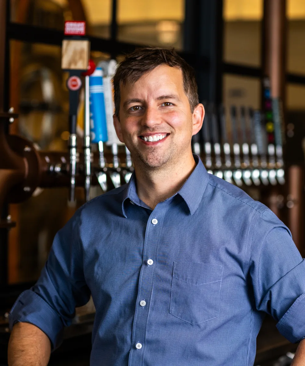 Smiling man in a blue checkered shirt standing in front of a row of beer taps in a brewery or bar setting.