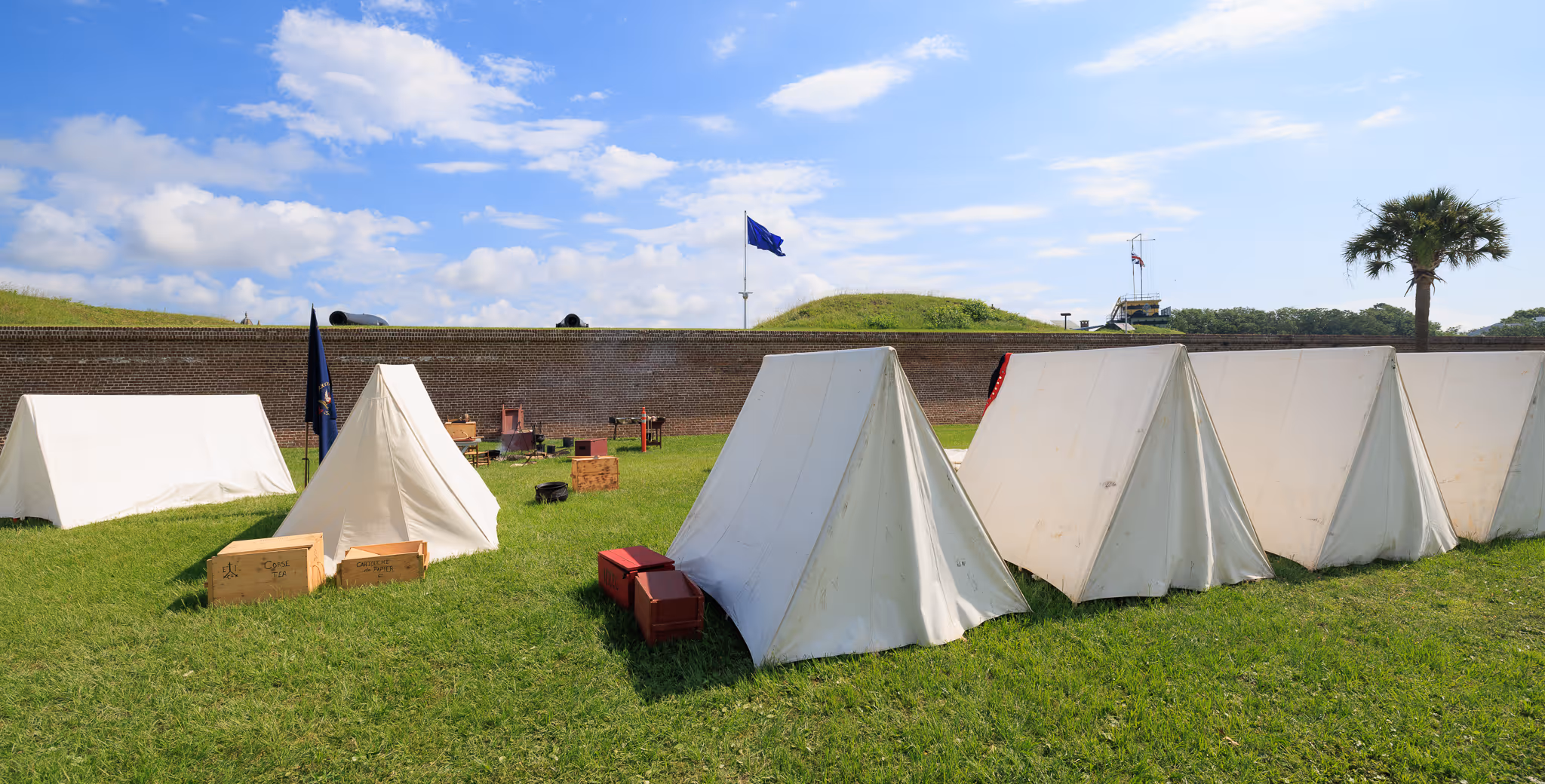Row of white canvas tents on green grass with wooden crates, a brick wall, and flags in the background under a partly cloudy sky.