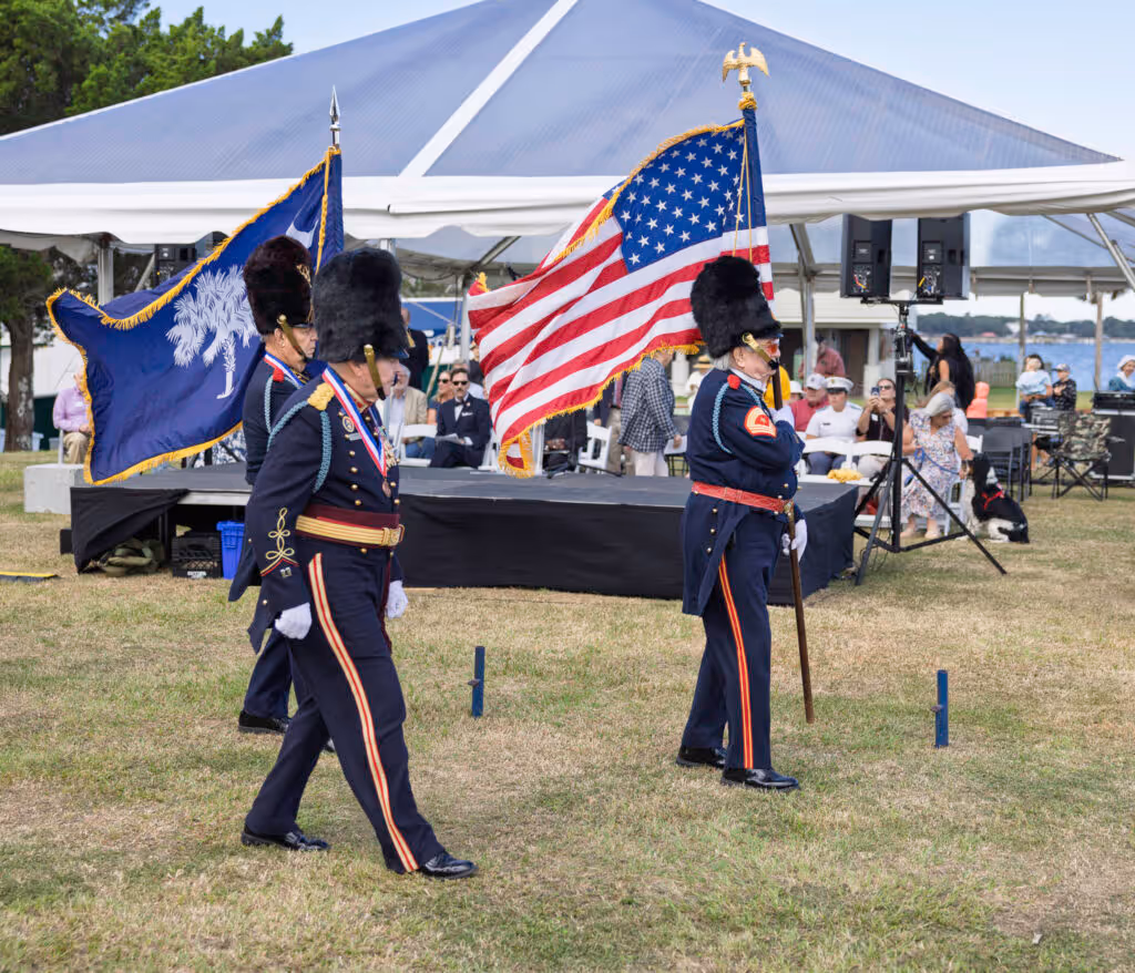 Three uniformed color guard members marching on grass carrying American and South Carolina flags with people seated under a white tent in the background.