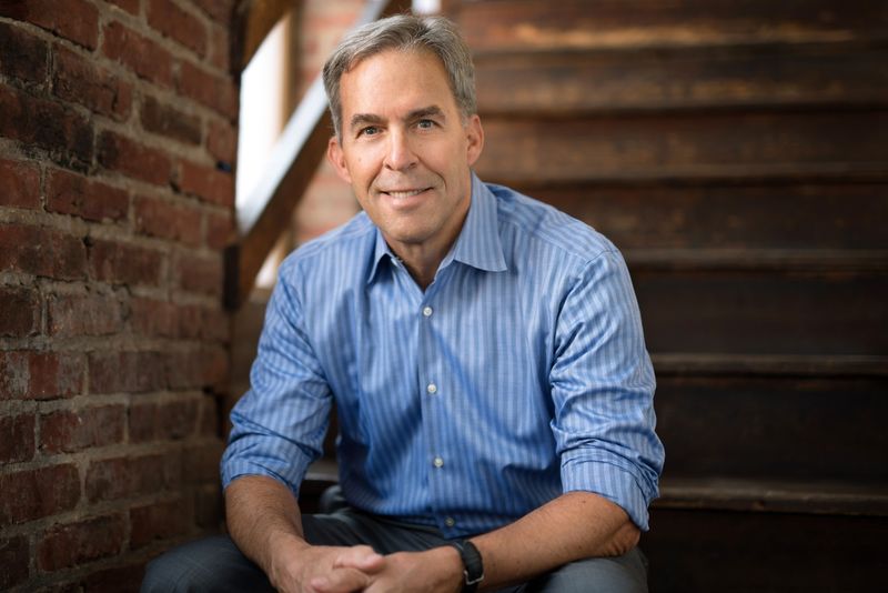 Middle-aged man in a blue striped shirt sitting on wooden stairs by a brick wall, smiling.