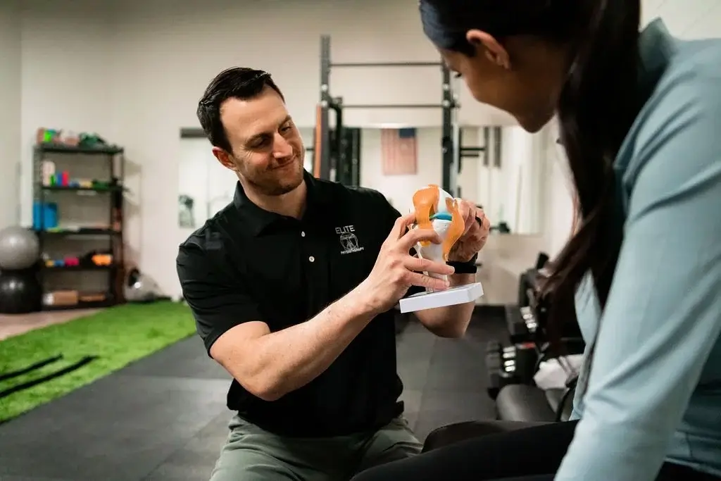 Physical therapist explaining a knee joint model to a female patient in a therapy gym.