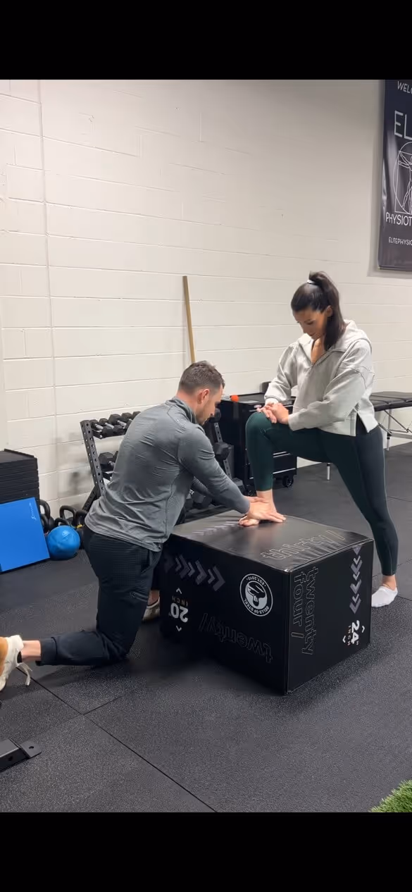 Man kneeling on one knee assisting a woman stretching her ankle on a workout box in a gym setting.