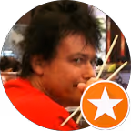 Volto di una persona con capelli scuri e maglietta rossa, con badge arancione con stella bianca.