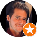 Ritratto di un uomo sorridente con capelli scuri e un distintivo arancione stella in basso a destra.