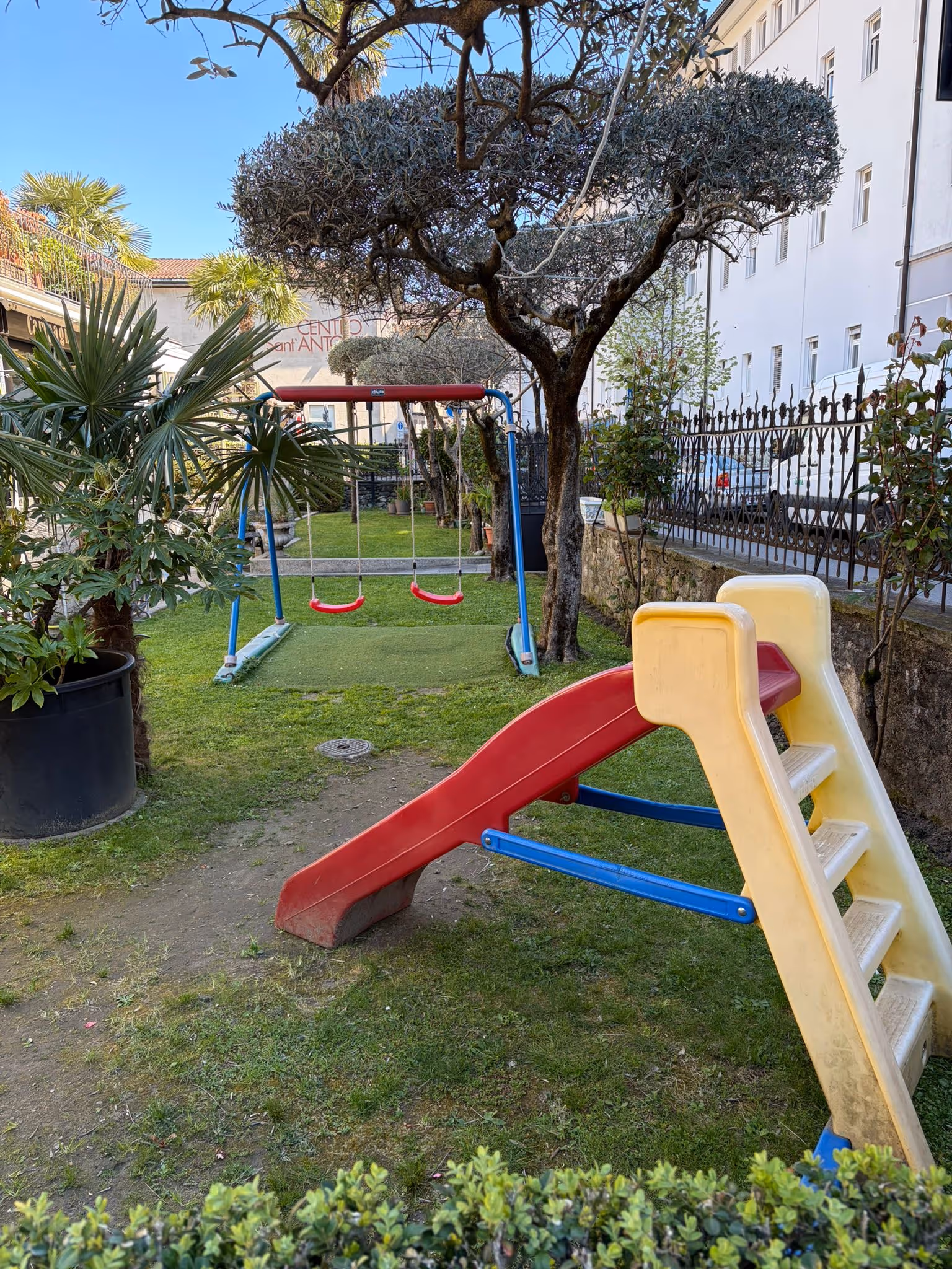 Scivolo e altalene in un piccolo parco giochi verde con alberi e cespugli attorno.