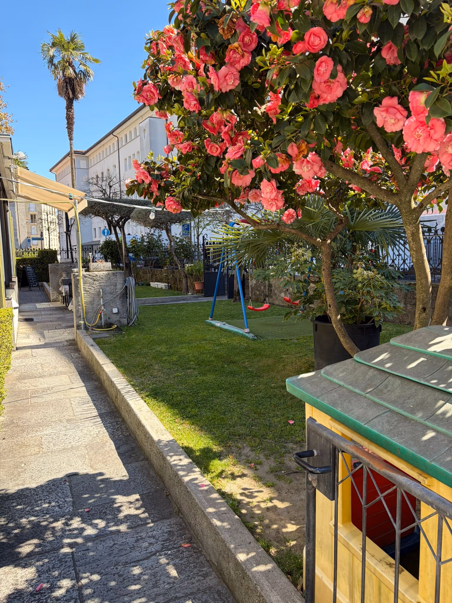 Giardino con fiori rosa, altalena rossa e casa gioco gialla vicino a un vialetto lastricato.