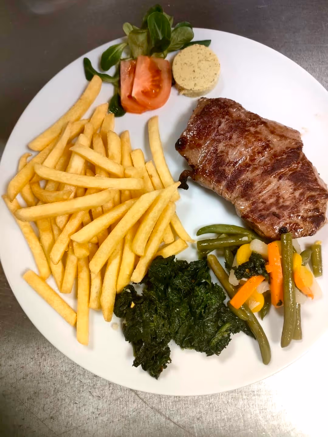 Piatto con bistecca, patatine fritte, spinaci, verdure miste, pomodori e burro alle erbe.