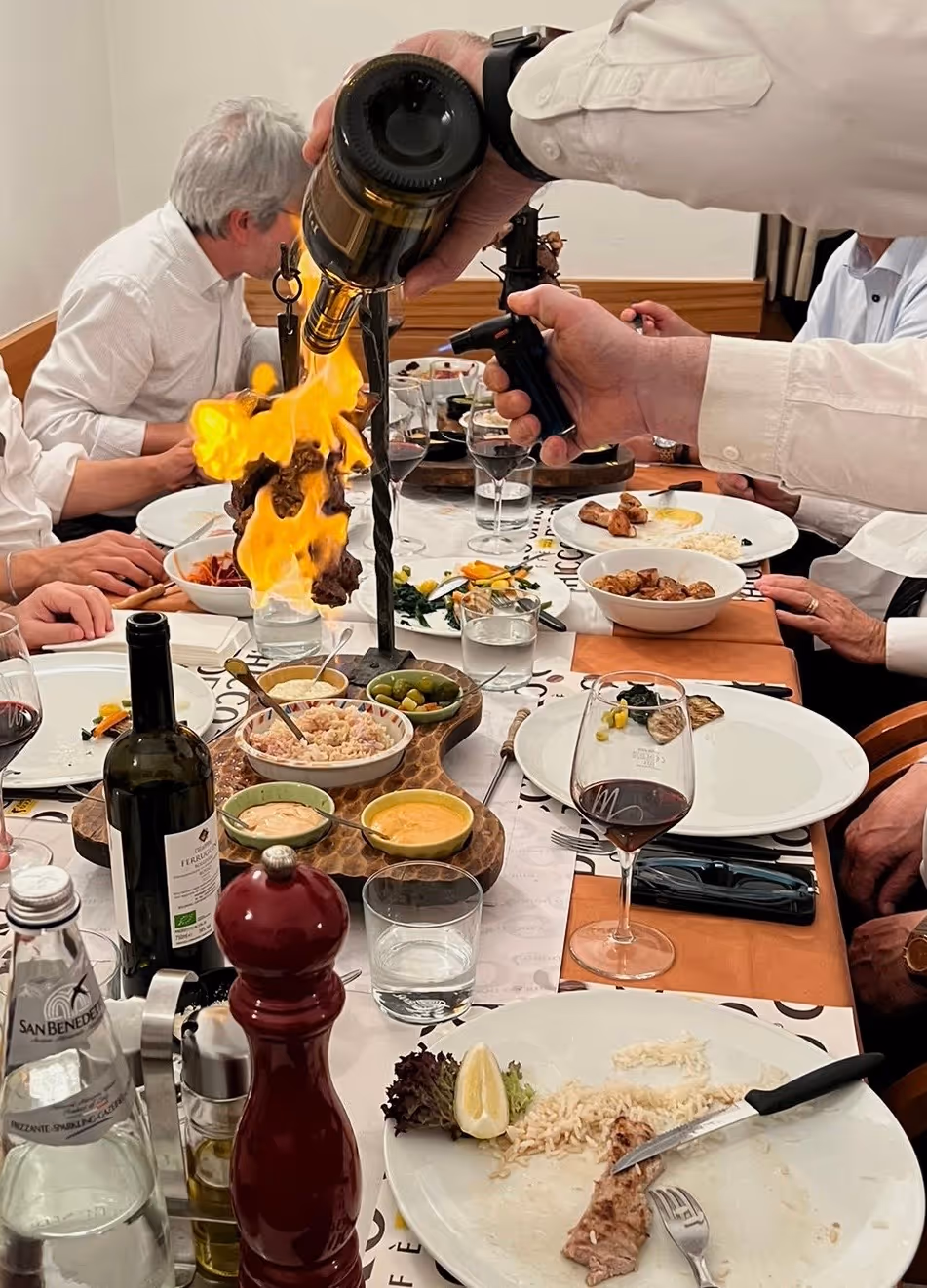 Persona versa liquido infiammabile su carne in fiamme a tavola con piatti e bicchieri di vino.