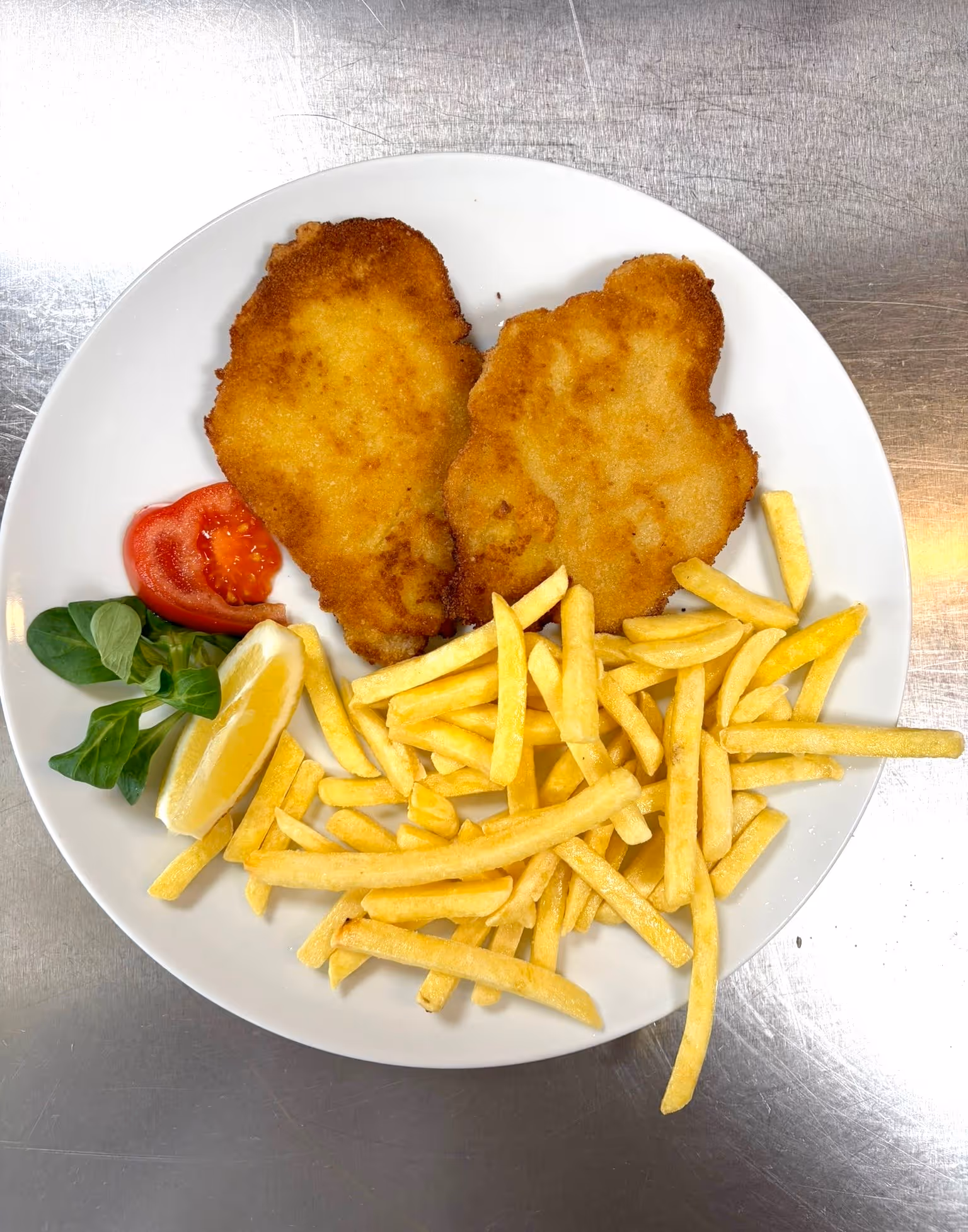 Petto di pollo impanato con patatine fritte, fetta di limone, pomodoro e insalata su piatto bianco.