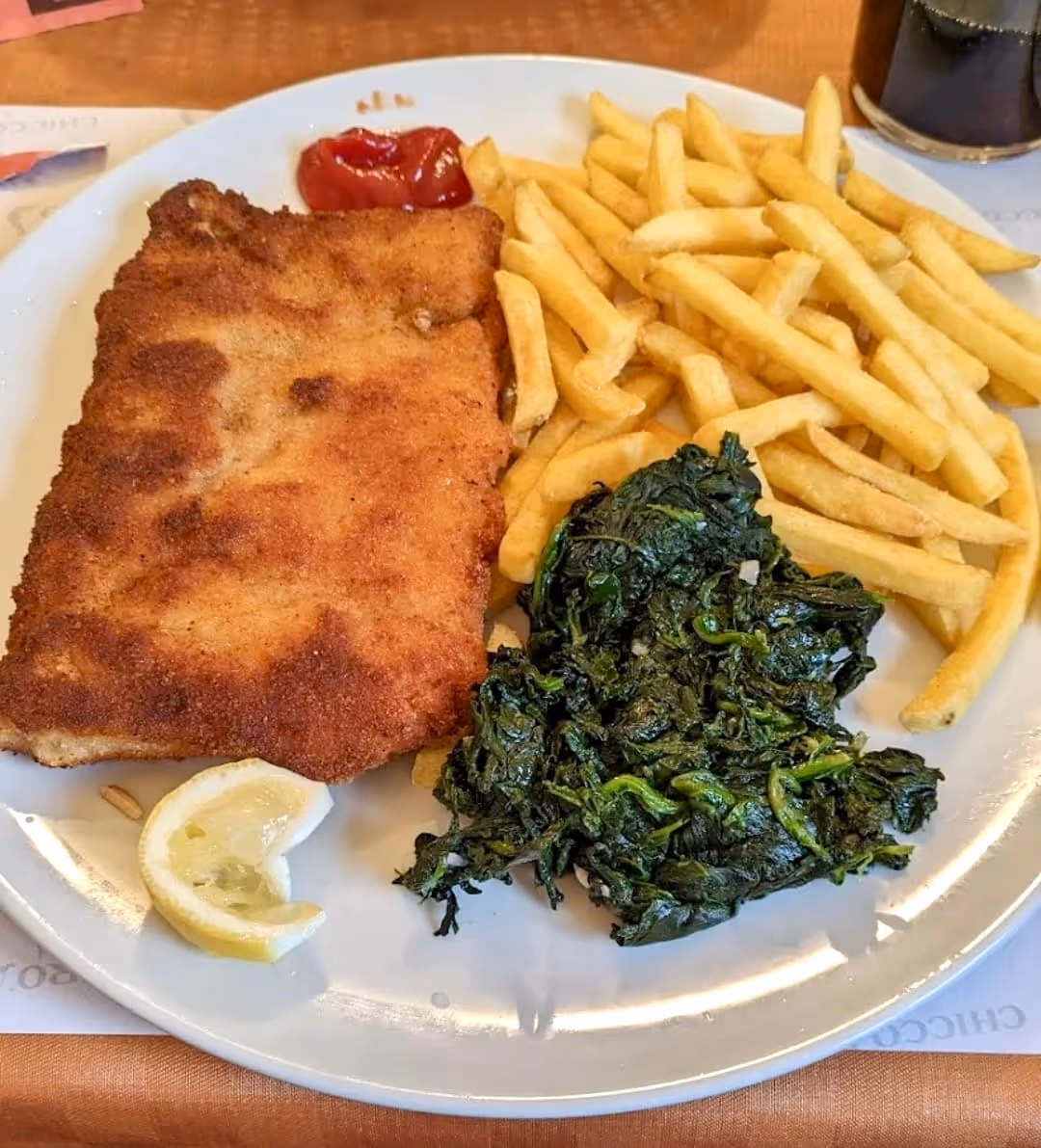 Cotoletta fritta con patatine fritte, spinaci saltati, fetta di limone e ketchup su un piatto bianco.
