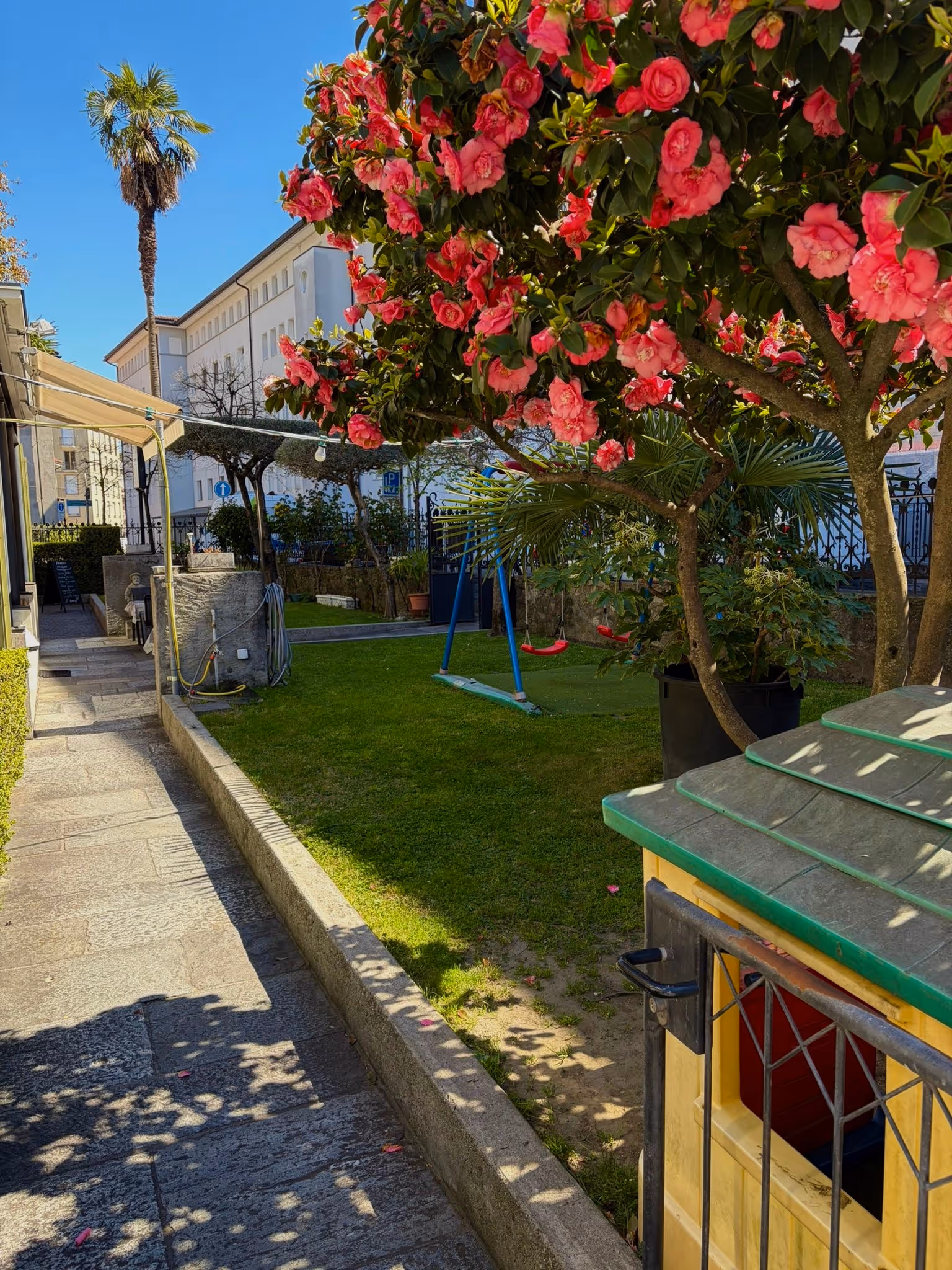 Giardino urbano con siepi, fiori rosa, altalena rossa e giochi per bambini sotto cielo azzurro.