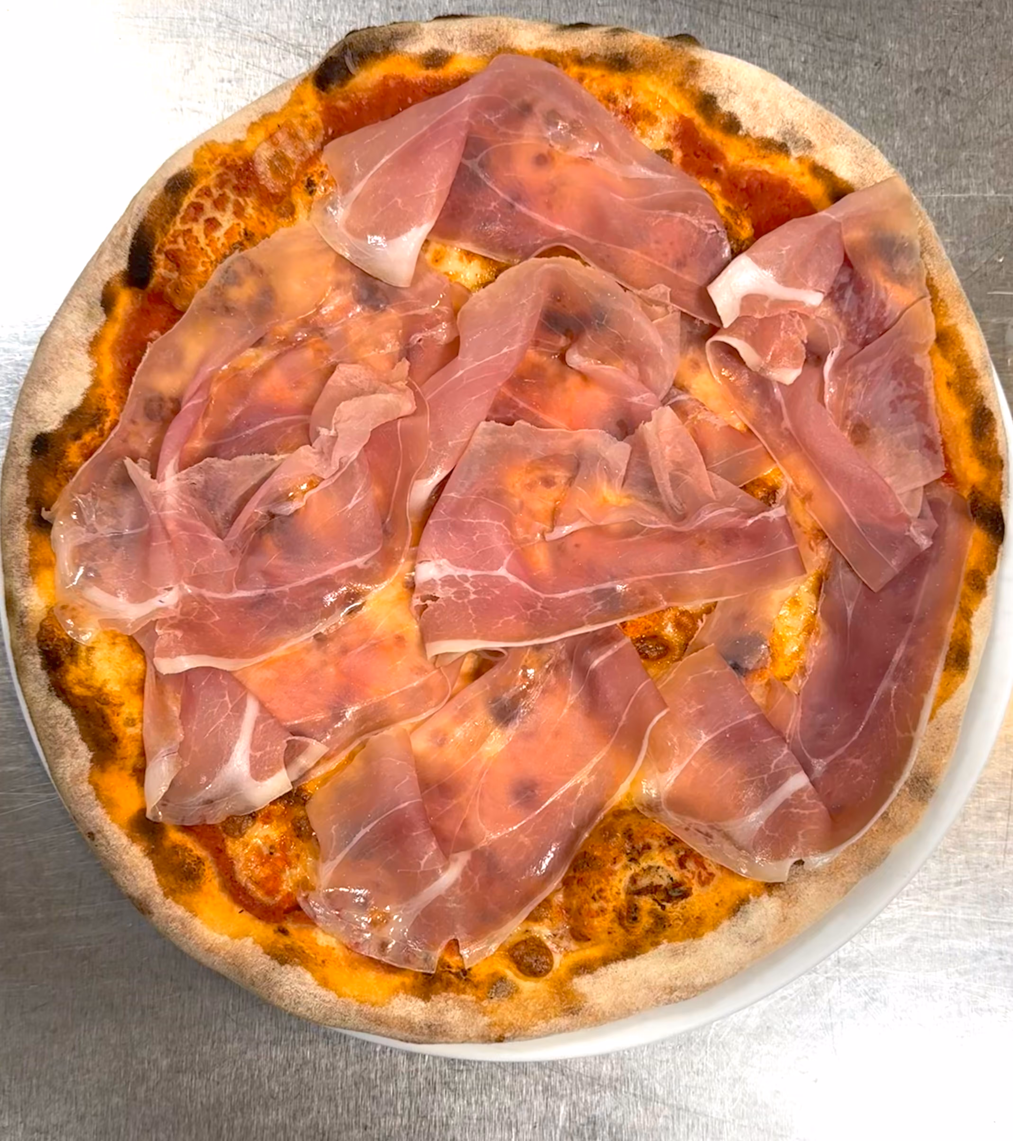 Pizza con prosciutto crudo su crosta dorata e salsa di pomodoro, servita su un piatto bianco.