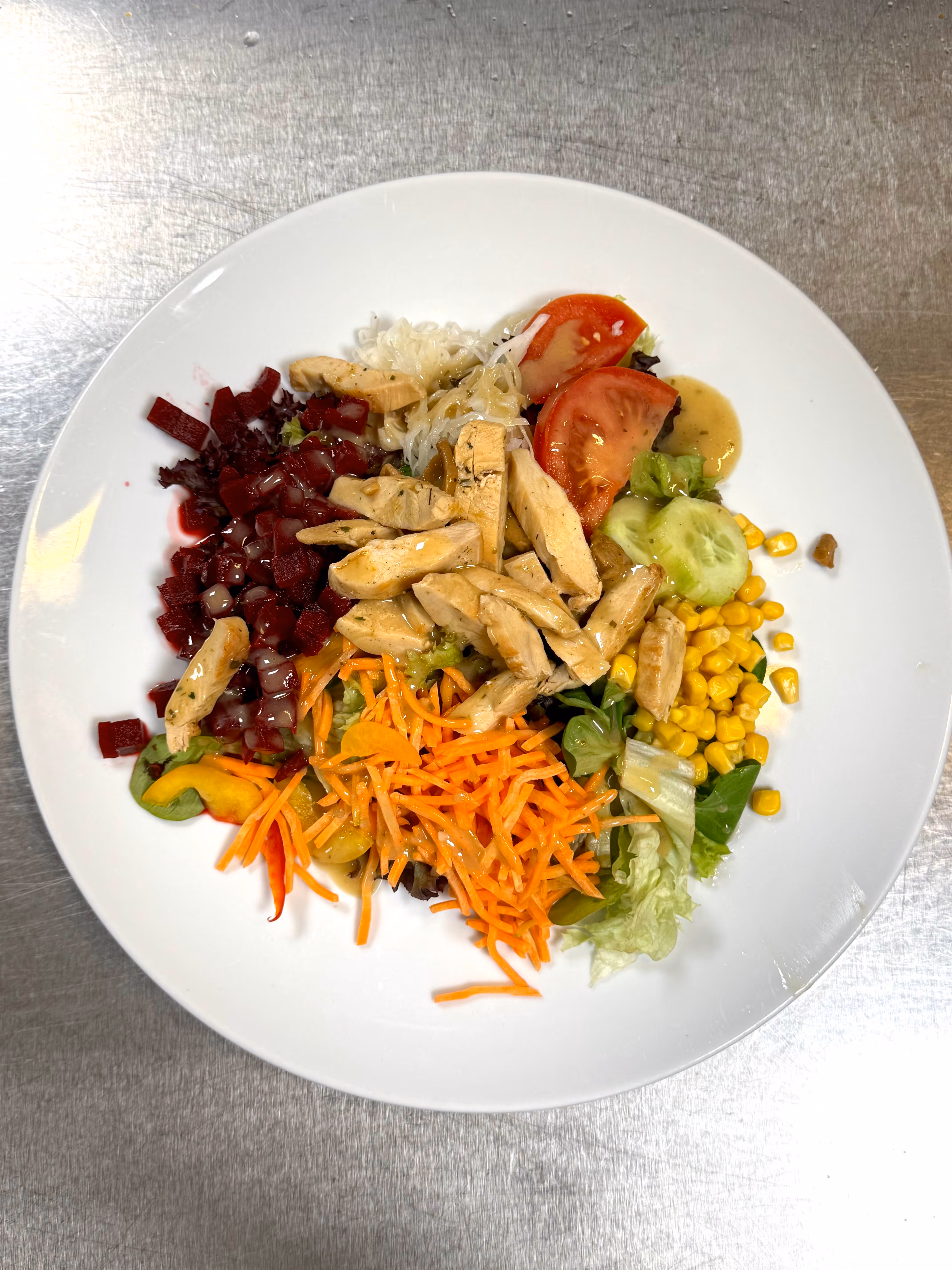 Insalata con pollo a fette, pomodoro, cetriolo, mais, carote, barbabietole e lattuga su piatto bianco.