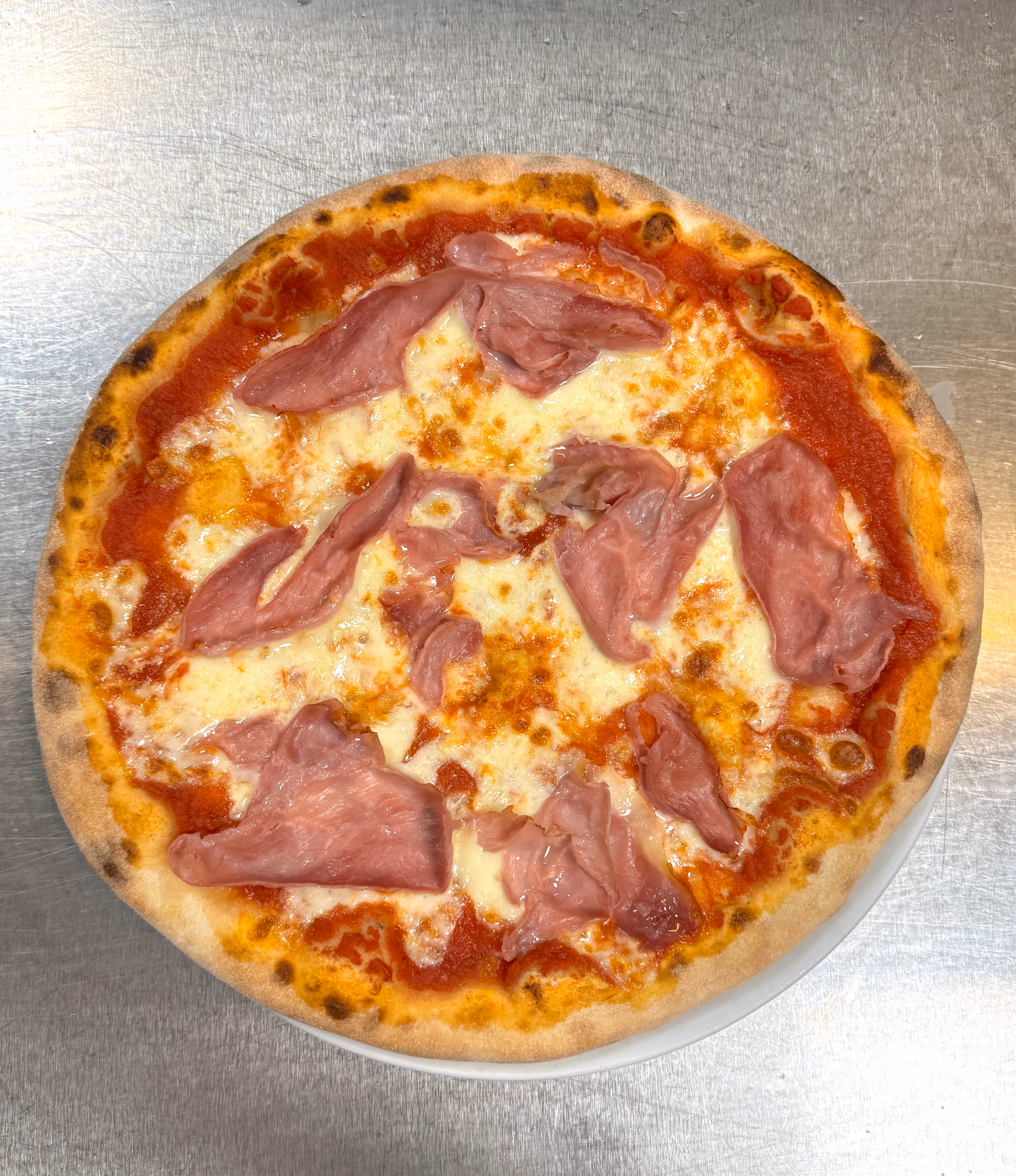 Pizza con salsa di pomodoro, mozzarella fusa e fette di prosciutto cotto su un fondo di acciaio.