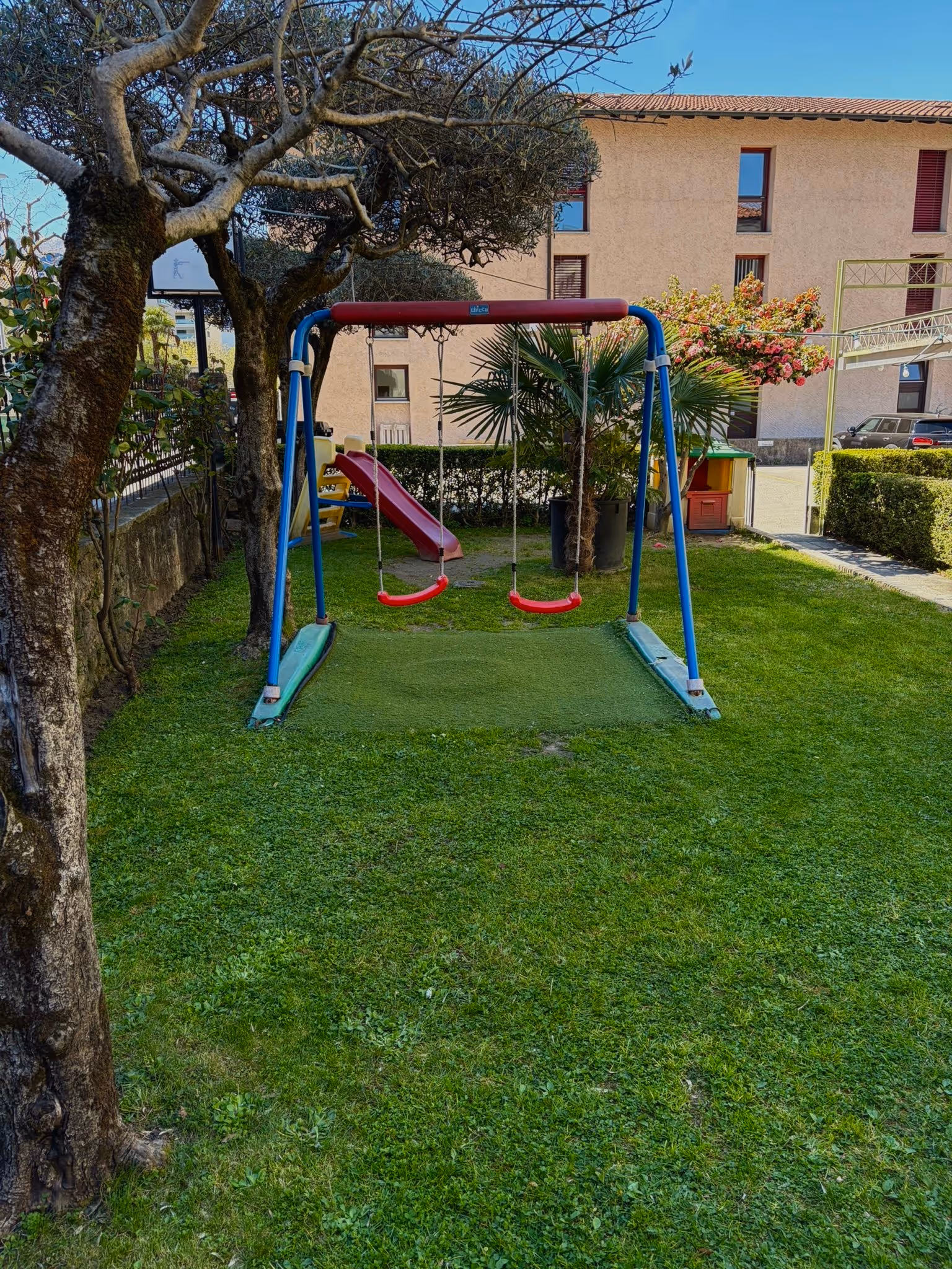 Parco giochi con altalena a due posti rossi, scivolo rosso e area verde con alberi.