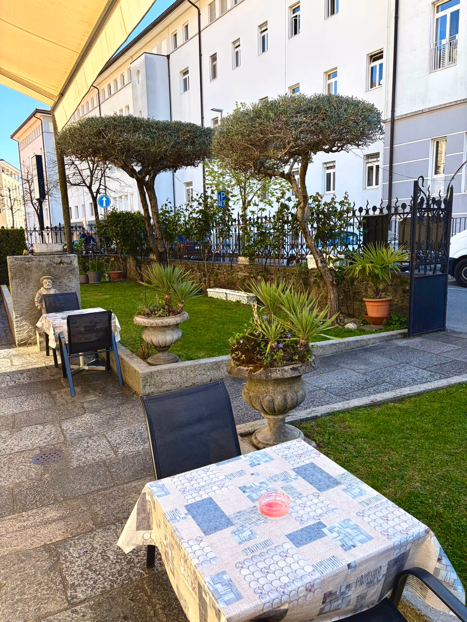 Tavoli con tovaglie e sedie nere in un giardino con alberi potati e vaso di pietra con piante.