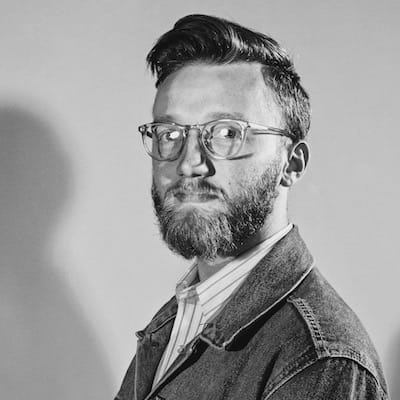 Bearded man wearing clear glasses and a denim jacket looking slightly to the side in grayscale photo.