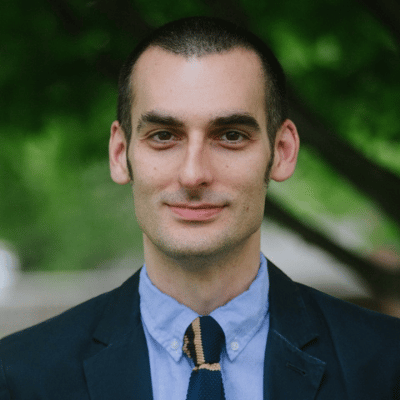 Man with short dark hair wearing a dark suit jacket, blue shirt, and striped tie, outdoors with trees behind.