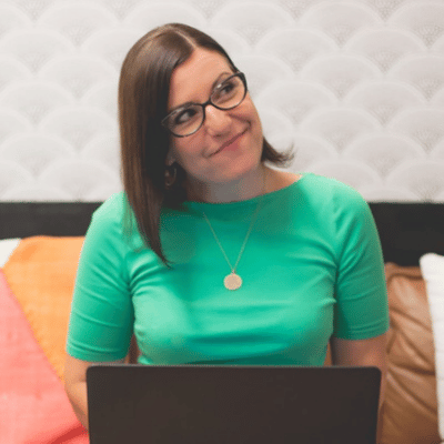 Woman with glasses in a green shirt smiling while sitting on a couch working on a laptop.