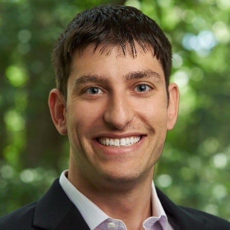 Smiling man with short dark hair in a suit jacket and white shirt against a green blurred background.