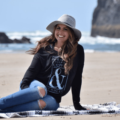 Smiling woman in a gray hat and black sweatshirt sitting on a blanket at the beach with ocean waves behind.