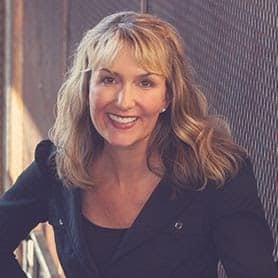 Smiling woman with wavy blonde hair wearing a black top and jacket in front of a textured wall.